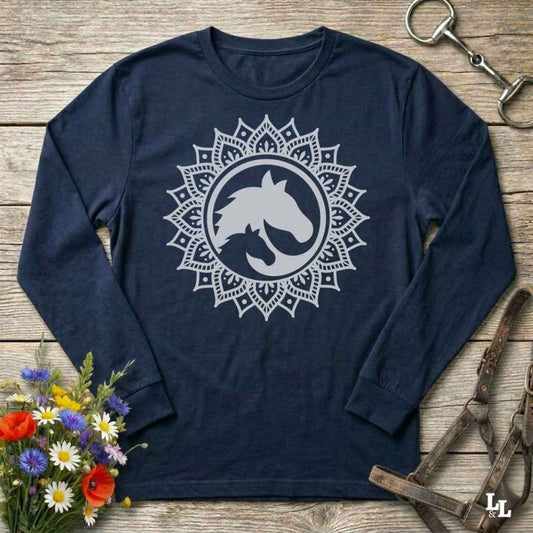 A long-sleeve crewneck shirt in navy blue , featuring a white graphic of a horse and cat silhouette inside a circular mandala design. The shirt is displayed flat on a rustic wooden background with equestrian tack and a small bouquet of flowers.