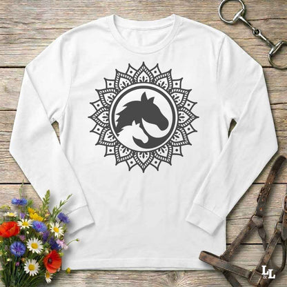 A long-sleeve crewneck shirt in white red, featuring a black graphic of a horse and cat silhouette inside a circular mandala design. The shirt is displayed flat on a rustic wooden background with equestrian tack and a small bouquet of flowers.