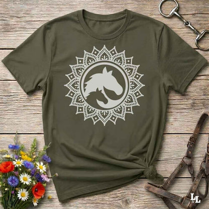 An olive green short-sleeve t-shirt with a white mandala graphic containing a horse silhouette, laid flat on a rustic wooden surface with decorative flowers and leather tack around it.