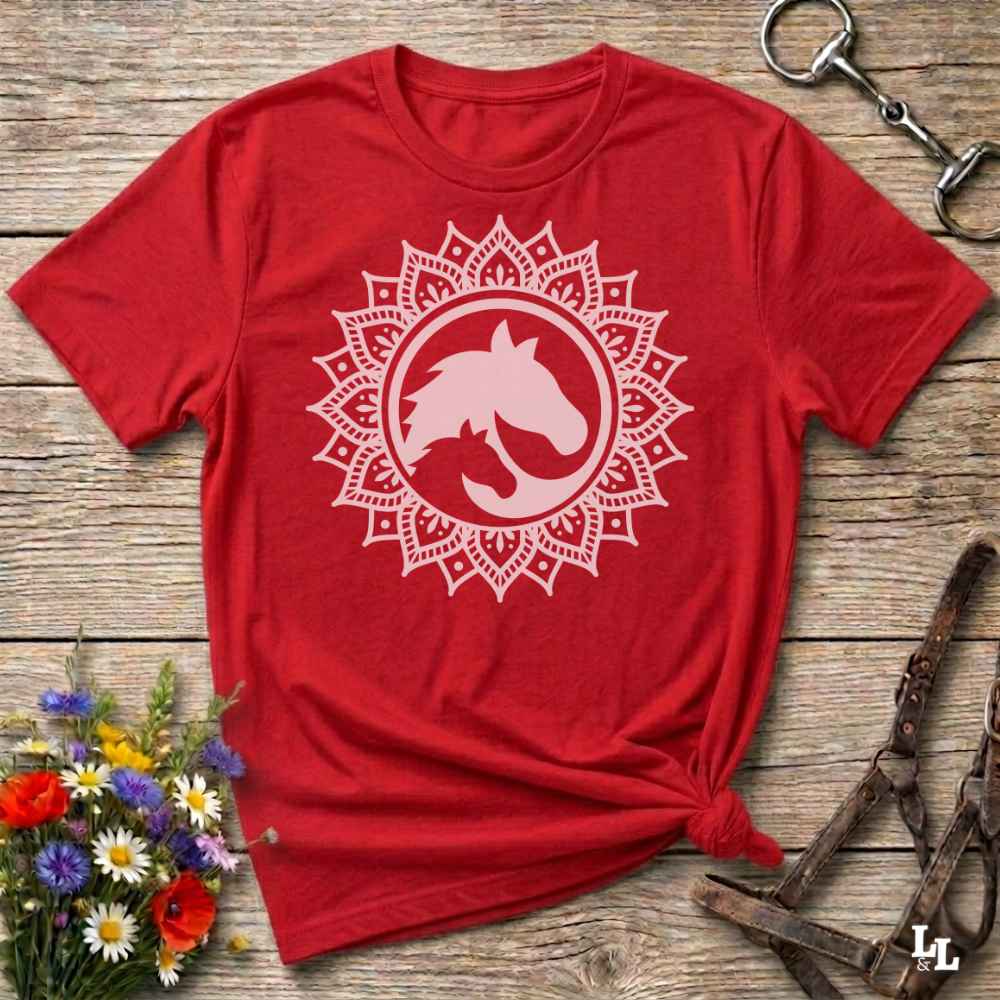 A red short-sleeve t-shirt with a white mandala-style horse graphic on the chest, styled flat on a rustic wooden surface with flowers and equestrian tack nearby.