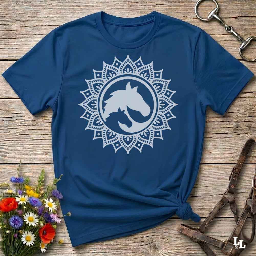 A blue short-sleeve t-shirt with a white mandala-style horse graphic on the chest, styled flat on a rustic wooden surface with flowers and equestrian tack nearby.