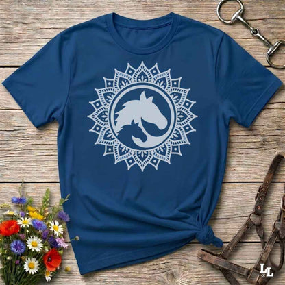 A blue short-sleeve t-shirt with a white mandala-style horse graphic on the chest, styled flat on a rustic wooden surface with flowers and equestrian tack nearby.