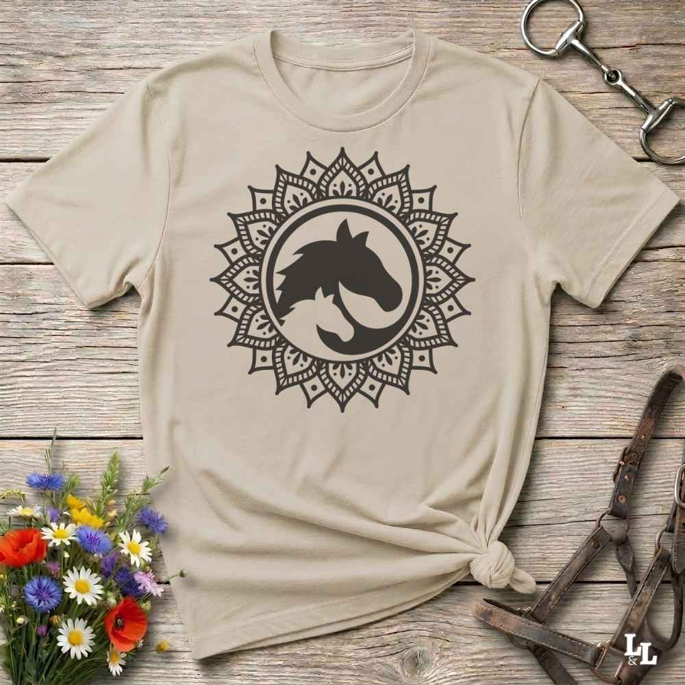 A beige short-sleeve t-shirt with a black horse head mandala graphic, styled in a flat-lay on a rustic wooden background with equestrian accessories and flowers.