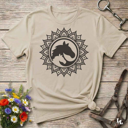 A beige short-sleeve t-shirt with a black horse head mandala graphic, styled in a flat-lay on a rustic wooden background with equestrian accessories and flowers.