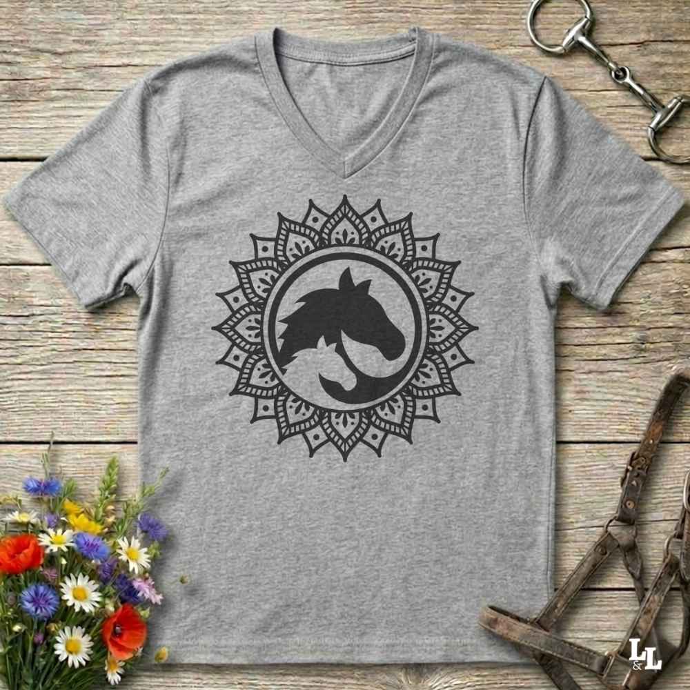 A heather gray V-neck t-shirt with a black horse silhouette inside a circular mandala design, laid flat on a rustic wooden surface with equestrian accessories and flowers as props.
