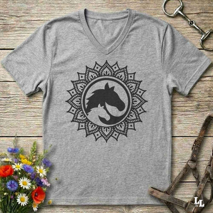 A heather gray V-neck t-shirt with a black horse silhouette inside a circular mandala design, laid flat on a rustic wooden surface with equestrian accessories and flowers as props.