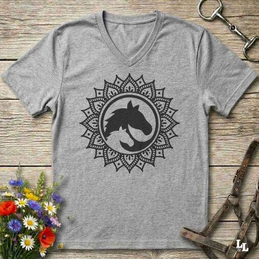 A heather gray V-neck t-shirt with a black horse silhouette inside a circular mandala design, laid flat on a rustic wooden surface with equestrian accessories and flowers as props.