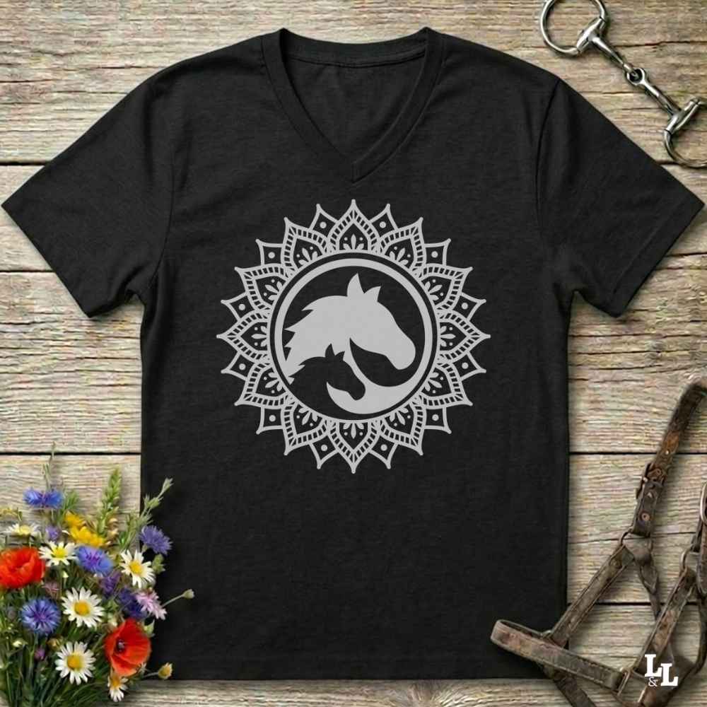 A black V-neck t-shirt with a white mandala-style horse graphic on the chest, laid flat on a rustic wooden surface with equestrian accessories and flowers around it.