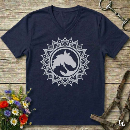 A navy blue V-neck t-shirt with a white mandala-style horse graphic on the chest, laid flat on a rustic wooden surface with equestrian accessories and flowers around it.