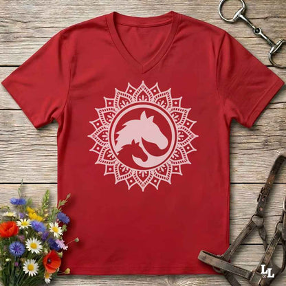 A red V-neck t-shirt with a white horse silhouette inside a mandala design, laid flat on a rustic wooden background with equestrian accessories and flowers.