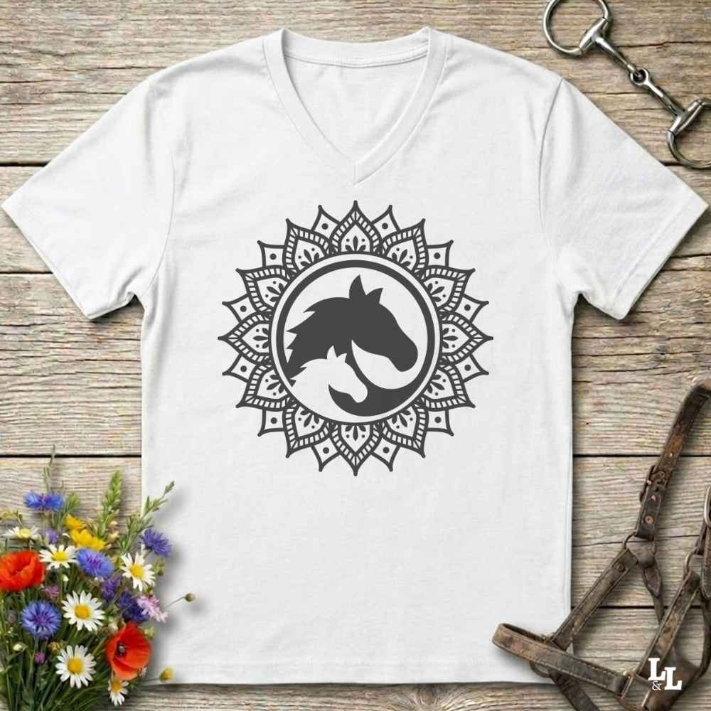 A white V-neck t-shirt with a black horse silhouette inside a circular mandala design, displayed on a rustic wooden background with equestrian and floral props.