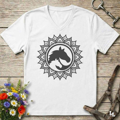 A white V-neck t-shirt with a black horse silhouette inside a circular mandala design, displayed on a rustic wooden background with equestrian and floral props.