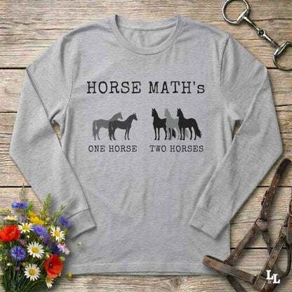 A light gray long-sleeve t-shirt with a 'Horse Math's' graphic printed on the chest, laid flat on a rustic wooden surface with equestrian-themed props like a halter, leather bridle, and a small bouquet of wildflowers.