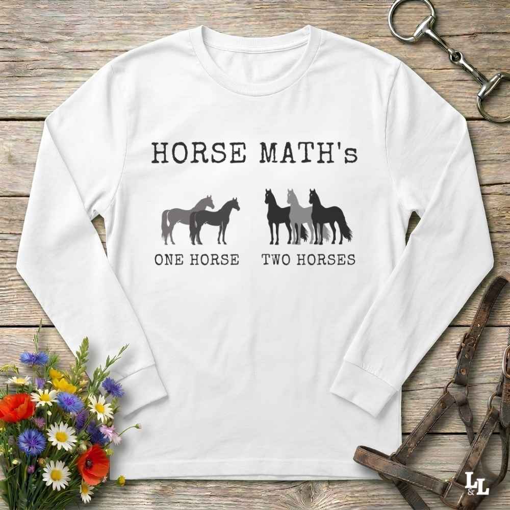 A white long-sleeve t-shirt with a black graphic print reading 'HORSE MATH'S' and 'ONE HORSE TWO HORSES' with horse silhouettes, laid flat on a rustic wooden surface with decorative flowers and equestrian accessories.