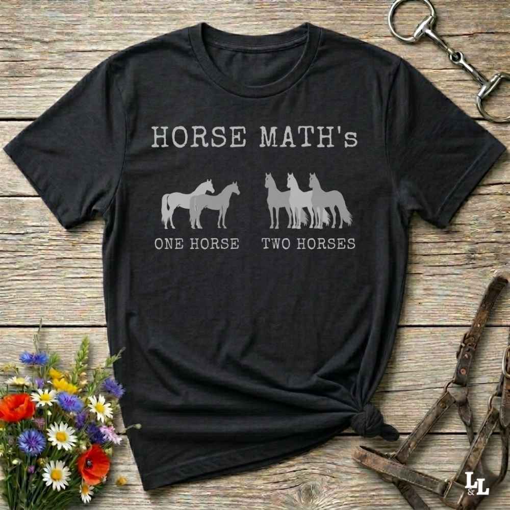 A black short-sleeve graphic T-shirt laid flat on a rustic wooden surface, with white text reading 'HORSE MATH'S' and horse silhouettes above the phrases 'ONE HORSE' and 'TWO HORSES.' Decorative flowers and equestrian straps appear around the shirt, but no person or real environment is present, so this is Product/Packaging rather than Lifestyle.