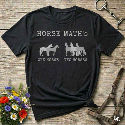 A black short-sleeve graphic T-shirt laid flat on a rustic wooden surface, with white text reading 'HORSE MATH'S' and horse silhouettes above the phrases 'ONE HORSE' and 'TWO HORSES.' Decorative flowers and equestrian straps appear around the shirt, but no person or real environment is present, so this is Product/Packaging rather than Lifestyle.