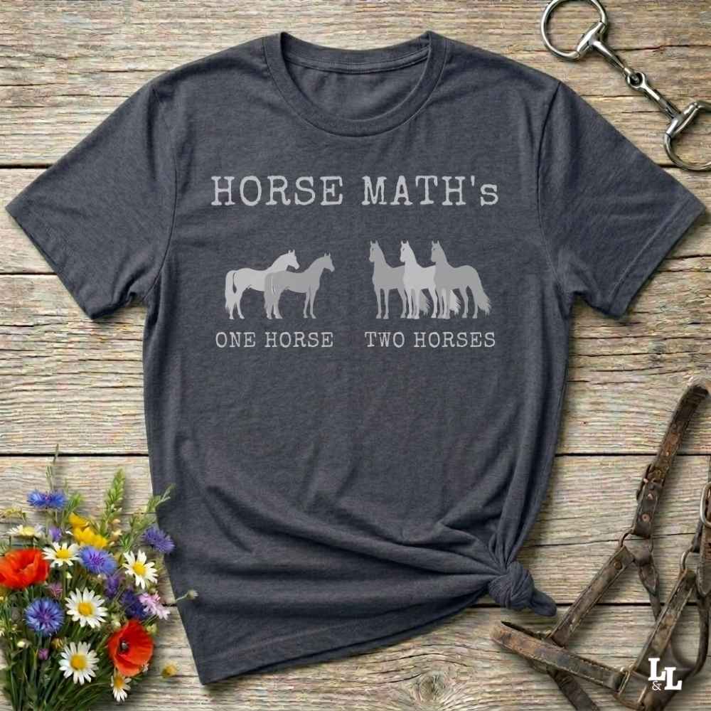 A dark heather gray graphic t-shirt laid flat on a rustic wooden surface, styled with a small bouquet of colorful wildflowers on the bottom left and horse tack on the top right. The t-shirt features a white graphic that reads 'HORSE MATH'S' above silhouettes of horses, with 'ONE HORSE' and 'TWO HORSES' printed below.