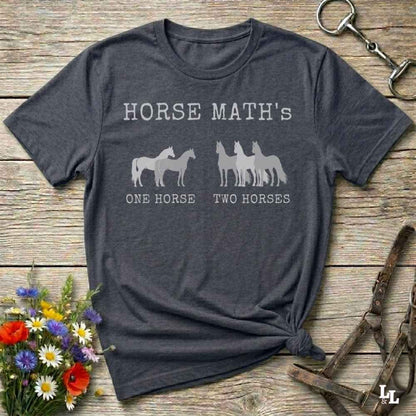 A dark heather gray graphic t-shirt laid flat on a rustic wooden surface, styled with a small bouquet of colorful wildflowers on the bottom left and horse tack on the top right. The t-shirt features a white graphic that reads 'HORSE MATH'S' above silhouettes of horses, with 'ONE HORSE' and 'TWO HORSES' printed below.