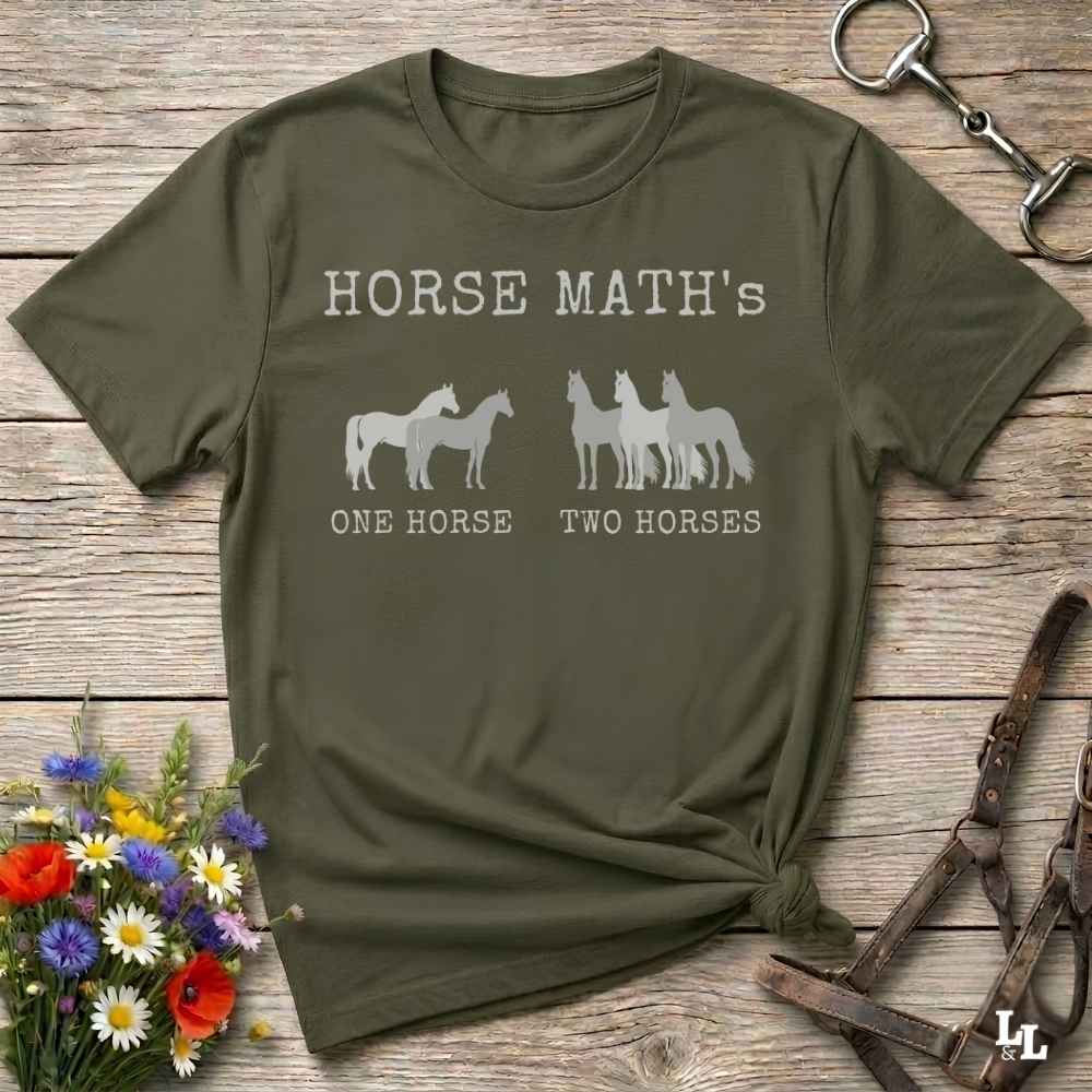 An olive green short-sleeve t-shirt with a white graphic print reading 'HORSE MATH'S' and 'ONE HORSE TWO HORSES' with horse silhouettes, styled on a rustic wooden background with equestrian accessories and flowers.