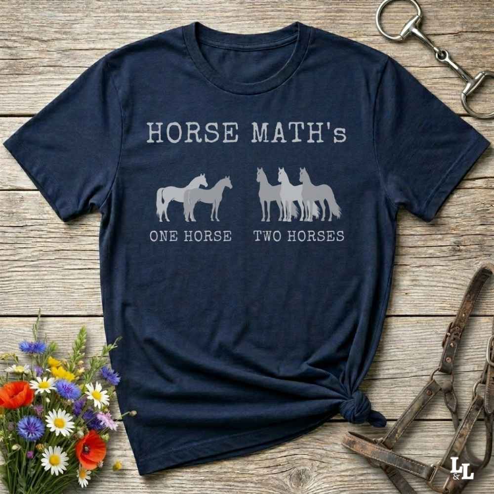 A navy blue graphic t-shirt laid flat on a rustic wooden surface, styled with flowers, a horse bit, and a leather halter. The shirt features a white graphic that reads 'HORSE MATH'S' above silhouettes of one and two horses, with 'ONE HORSE' and 'TWO HORSES' printed below.