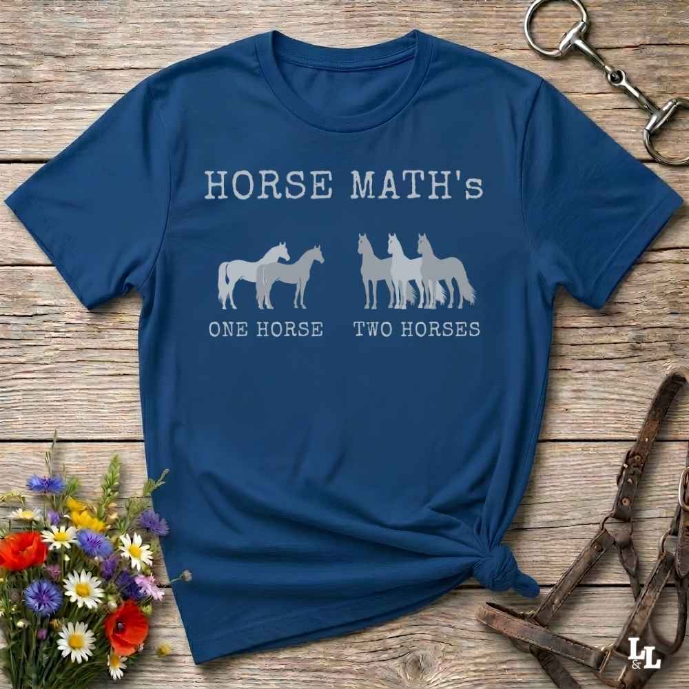 A navy blue graphic t-shirt laid flat on a rustic wooden surface, styled with a small bouquet of wildflowers and equestrian accessories. The shirt features the text 'HORSE MATH'S' in white, followed by white horse silhouettes, and the phrases 'ONE HORSE' and 'TWO HORSES' underneath.