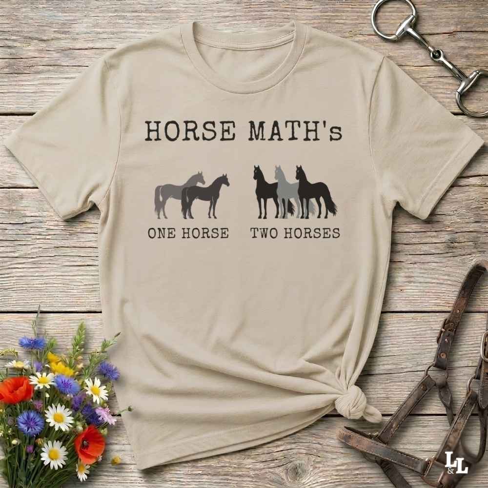 A beige  short-sleeve graphic T-shirt laid flat on a rustic wooden surface, with white text reading 'HORSE MATH'S' and horse silhouettes above the phrases 'ONE HORSE' and 'TWO HORSES.' Decorative flowers and horse tack are placed around the shirt as props.