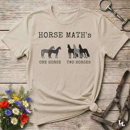 A beige  short-sleeve graphic T-shirt laid flat on a rustic wooden surface, with white text reading 'HORSE MATH'S' and horse silhouettes above the phrases 'ONE HORSE' and 'TWO HORSES.' Decorative flowers and horse tack are placed around the shirt as props.