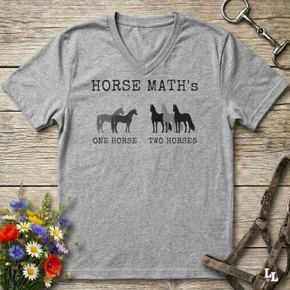 A heather gray V-neck t-shirt with a black graphic print reading 'HORSE MATH'S' above horse silhouettes and the phrases 'ONE HORSE' and 'TWO HORSES'. The shirt is laid flat on a rustic wood surface with equestrian accessories and flowers around it.