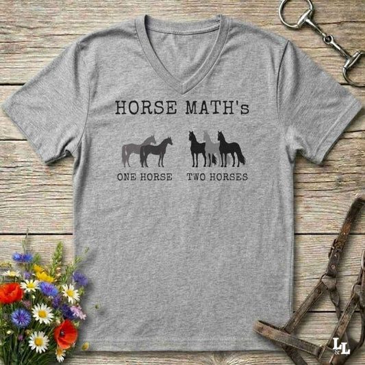 A heather gray V-neck t-shirt with a black graphic print reading 'HORSE MATH'S' above horse silhouettes and the phrases 'ONE HORSE' and 'TWO HORSES'. The shirt is laid flat on a rustic wood surface with equestrian accessories and flowers around it.