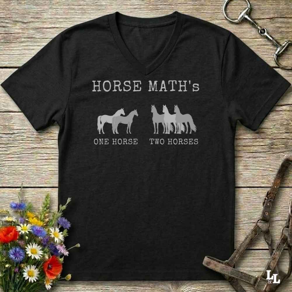 A black V-neck t-shirt with a gray graphic print reading 'HORSE MATH's' and 'ONE HORSE TWO HORSES' with horse silhouettes, displayed on a rustic wooden background with equestrian accessories and flowers.