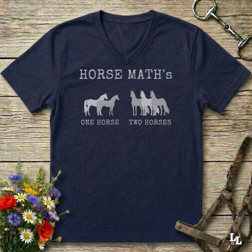 A navy blue V-neck t-shirt with a white graphic print reading 'HORSE MATH's' and 'ONE HORSE TWO HORSES' with horse silhouettes, laid flat on a rustic wooden surface with equestrian accessories and flowers around it.