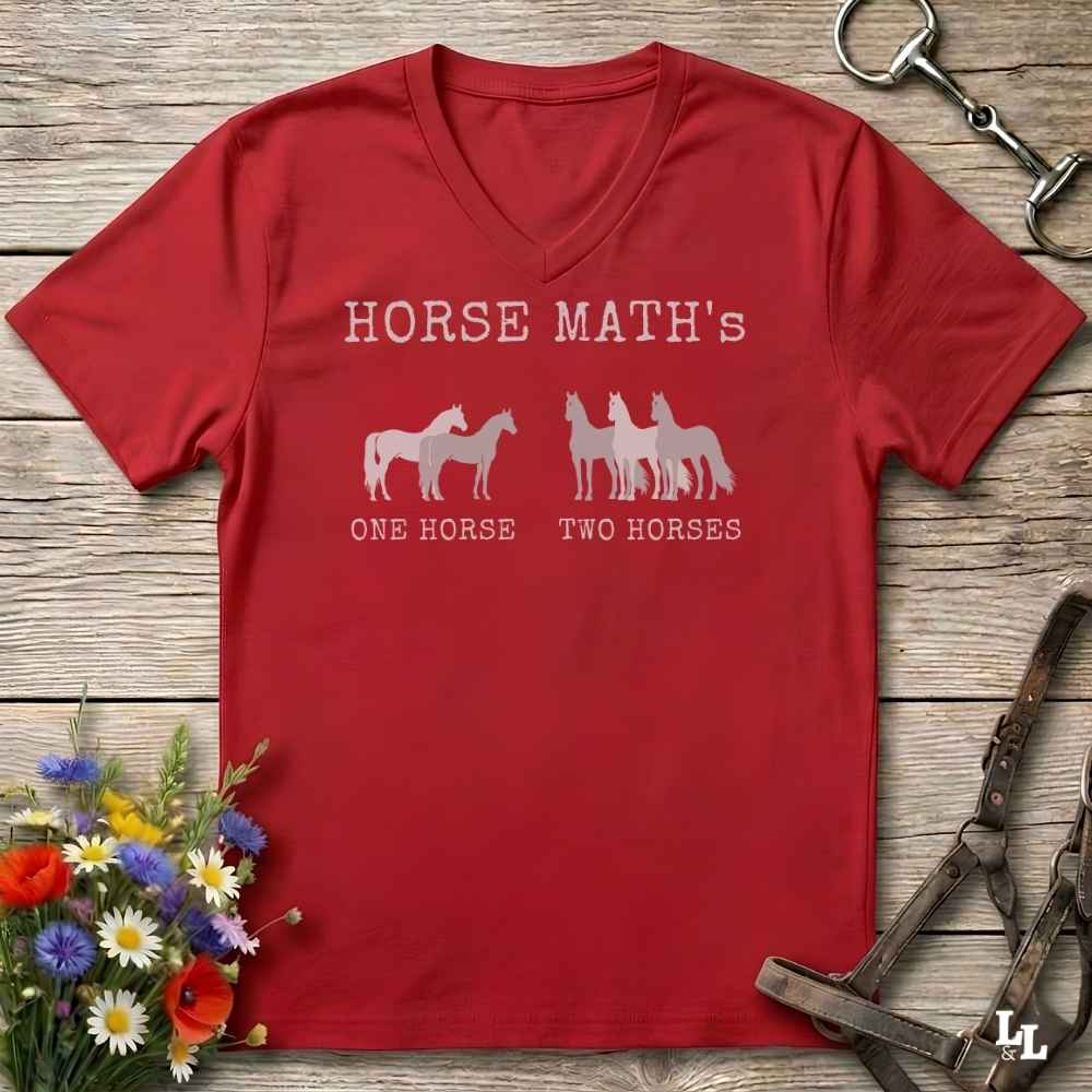 A red V-neck t-shirt laid flat on a rustic wooden surface, featuring a white graphic that reads 'HORSE MATH's' above horse silhouettes labeled 'ONE HORSE' and 'TWO HORSES'. The shirt is styled with flowers, a horse bit, and a halter.