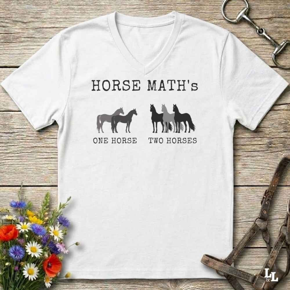 A white V-neck t-shirt with a black graphic print reading 'HORSE MATH's' and 'ONE HORSE TWO HORSES' with silhouettes of horses, laid flat on a rustic wooden surface with equestrian accessories and flowers around it.