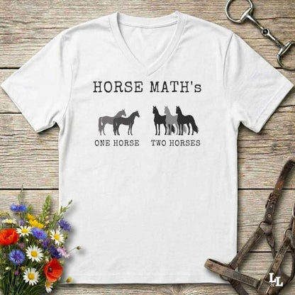 A white V-neck t-shirt with a black graphic print reading 'HORSE MATH's' and 'ONE HORSE TWO HORSES' with silhouettes of horses, laid flat on a rustic wooden surface with equestrian accessories and flowers around it.