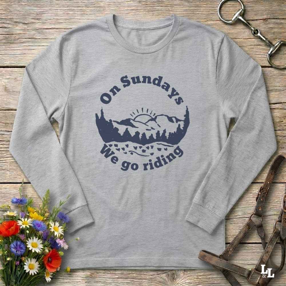 A light heather gray long-sleeve t-shirt with a navy blue graphic reading 'On Sundays We go riding' around a mountain and tree silhouette. The shirt is laid flat on a rustic wooden surface with decorative equestrian and floral props.
