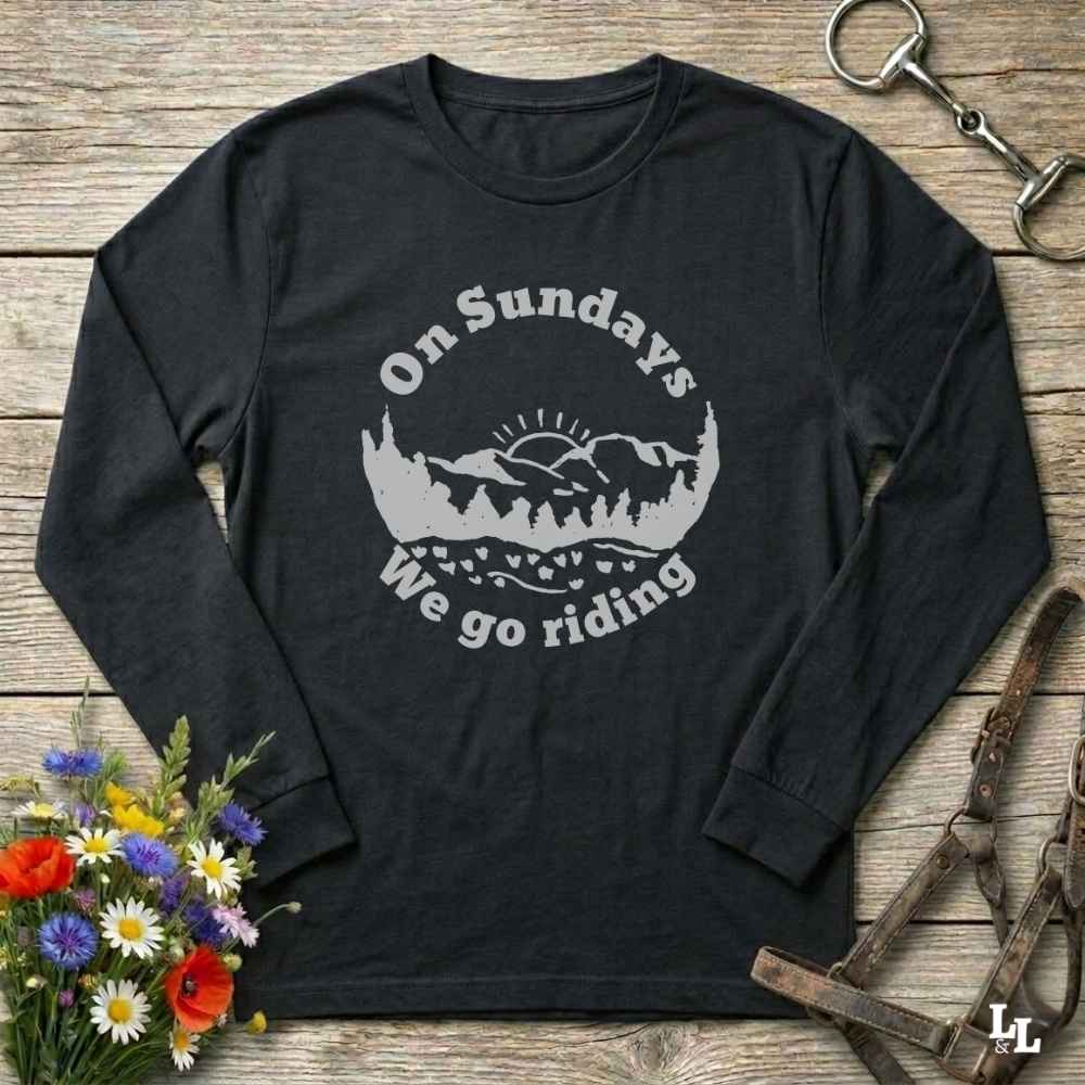 A black long-sleeve t-shirt with a gray graphic print reading 'On Sundays We go riding' around a mountain and sunset illustration, laid flat on a rustic wooden surface with flowers, a horse bit, and a leather strap nearby.