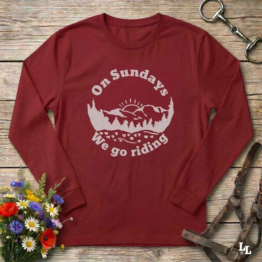 A long-sleeve crewneck t-shirt in maroon red with a white graphic print that reads 'On Sundays We go riding' surrounding a mountain and trees illustration. The shirt is laid flat on a rustic wooden background decorated with a small bouquet of flowers and equestrian tack.