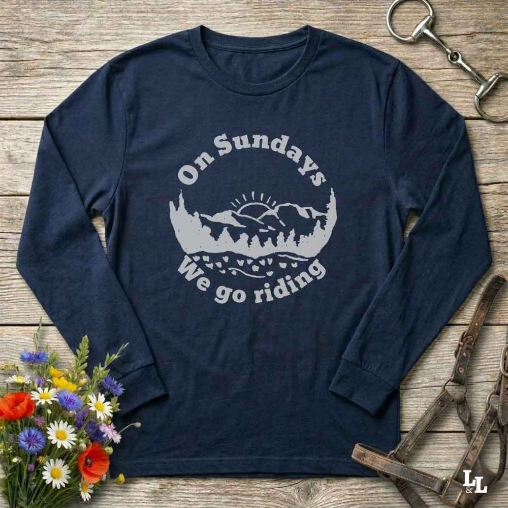 A navy blue long-sleeve t-shirt with a white graphic print that reads 'On Sundays We go riding' around a mountain and trees silhouette. The shirt is laid flat on a rustic wooden background with equestrian and floral props.