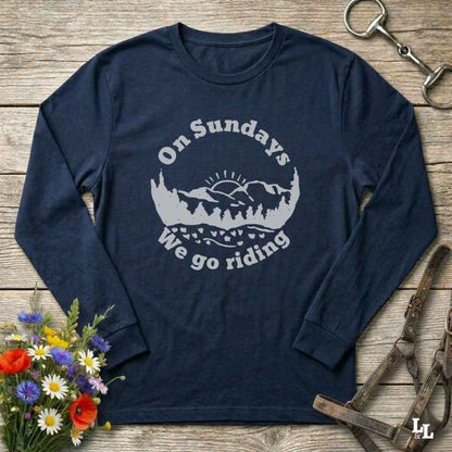 A navy blue long-sleeve t-shirt with a white graphic print that reads 'On Sundays We go riding' around a mountain and trees silhouette. The shirt is laid flat on a rustic wooden background with equestrian and floral props.