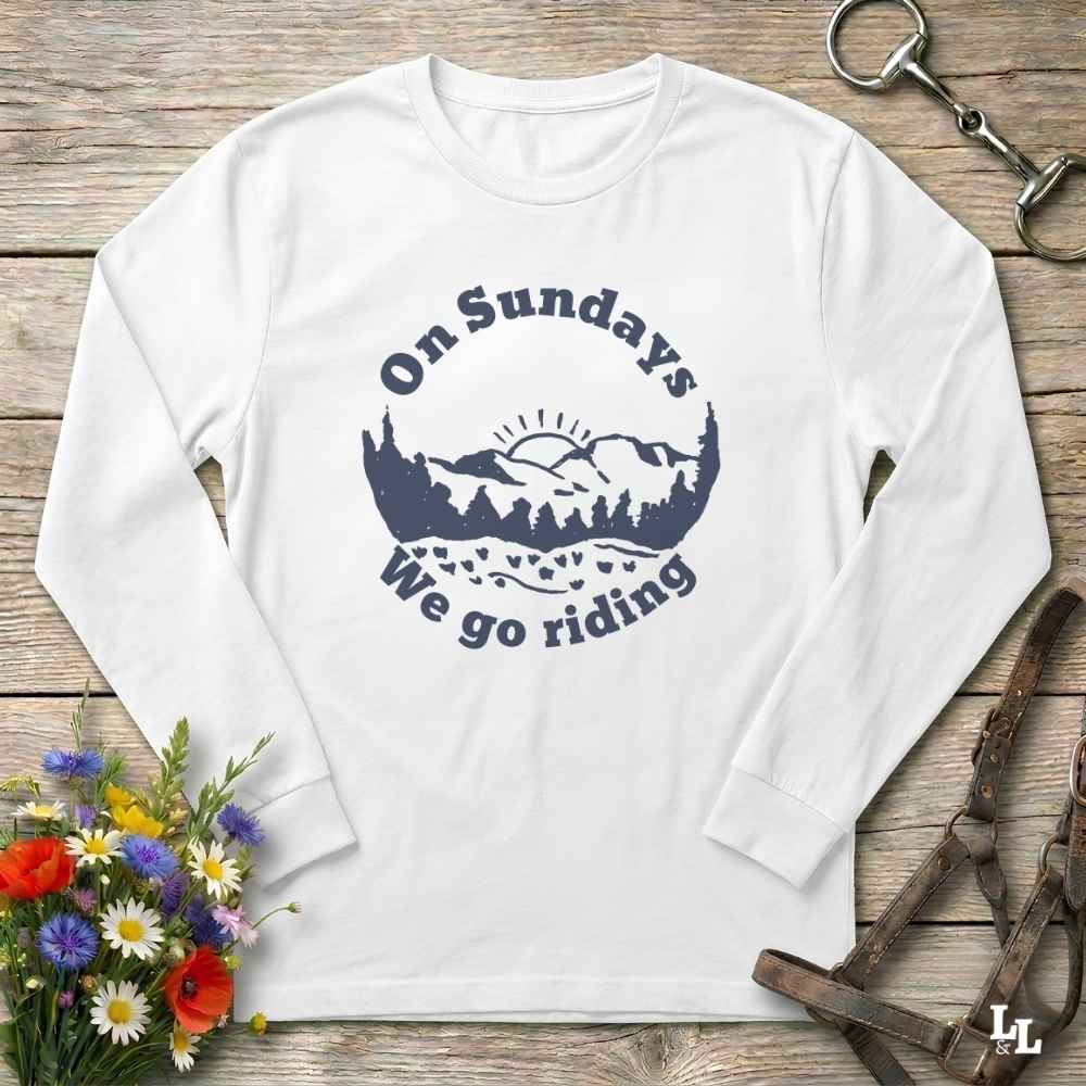 A white long-sleeve crewneck t-shirt with a navy blue graphic on the chest that reads 'On Sundays We go riding' surrounding a mountain and tree silhouette. The shirt is laid flat on a rustic wooden background with decorative equestrian and floral props.