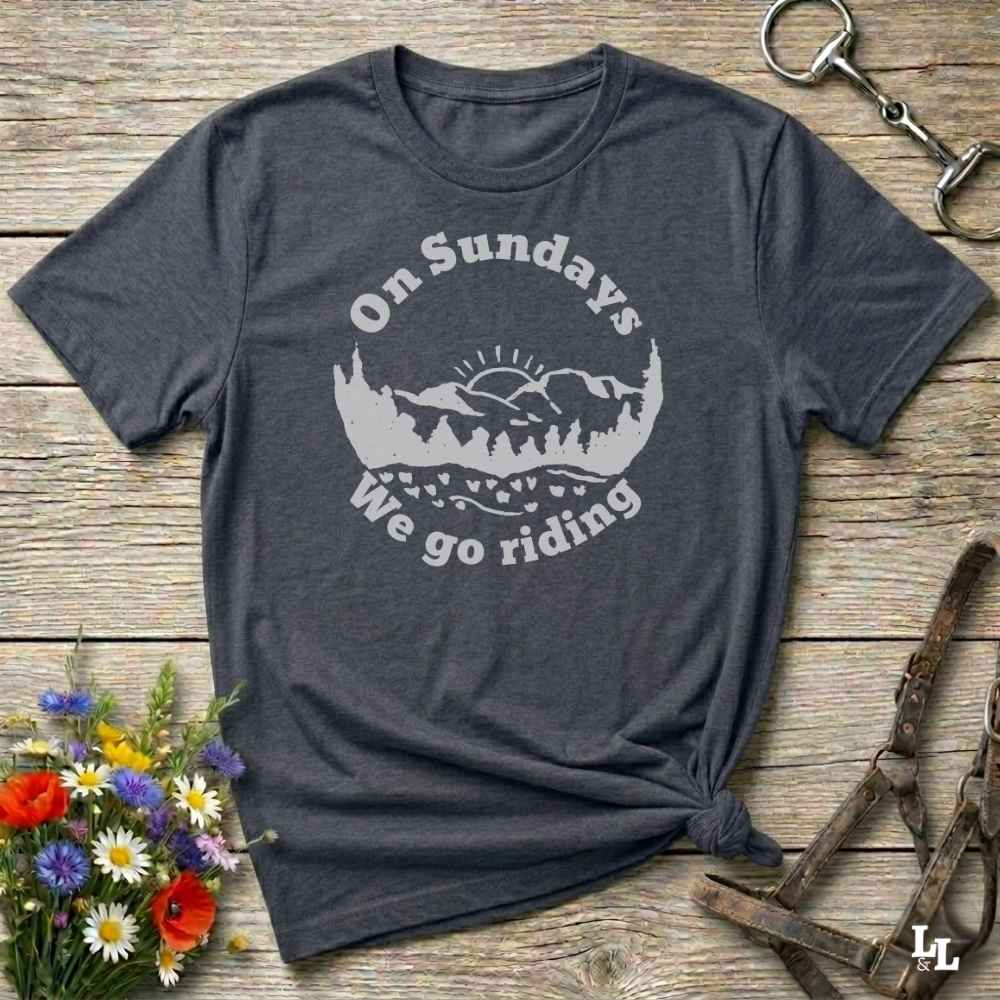 A dark heather gray t-shirt with a white graphic print reading 'On Sundays We go riding' around a mountain and tree silhouette. The shirt is styled in a flat lay on a rustic wooden background with a flower bouquet, a horse bit, and a leather halter.