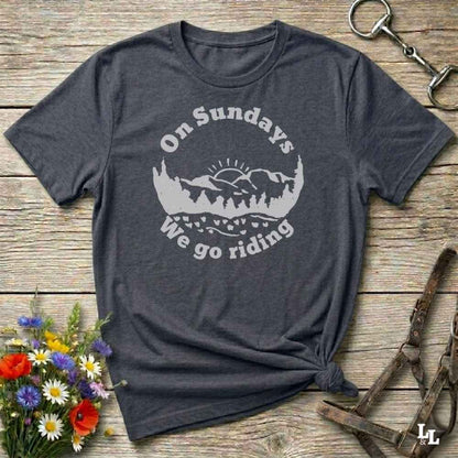 A dark heather gray t-shirt with a white graphic print reading 'On Sundays We go riding' around a mountain and tree silhouette. The shirt is styled in a flat lay on a rustic wooden background with a flower bouquet, a horse bit, and a leather halter.
