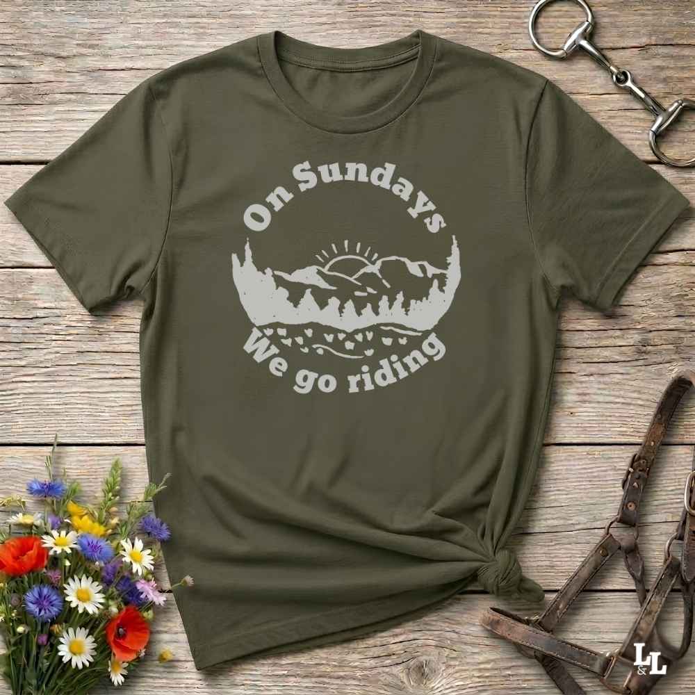 An olive green short-sleeve t-shirt with a white graphic reading 'On Sundays We go riding' around a mountain and tree silhouette, laid flat on a rustic wooden surface with flowers and horse tack props.
