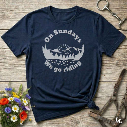 A navy blue short-sleeve t-shirt with a white graphic print reading 'On Sundays We go riding' around a mountain and tree silhouette. The shirt is styled in a flat-lay on a rustic wooden surface with decorative flowers, a horse bit, and a leather halter.