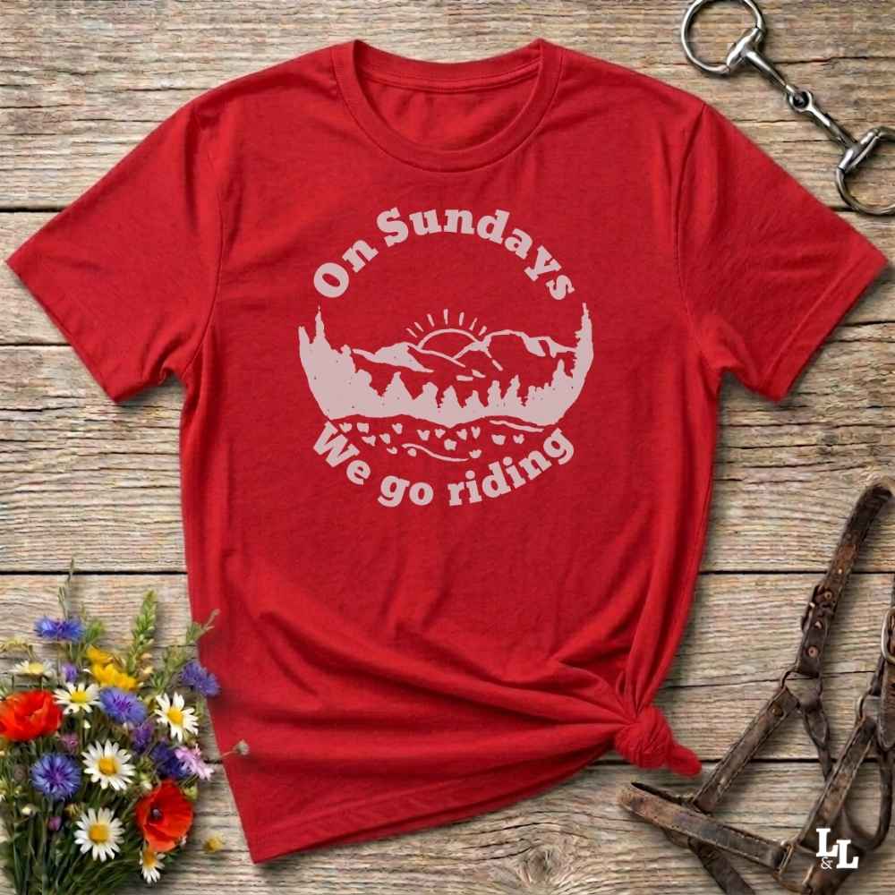 A red short-sleeve t-shirt with a white graphic reading 'On Sundays We go riding' and a mountain-and-tree illustration, styled flat on a rustic wood surface with flowers, a horse bit, and a leather bridle around it.