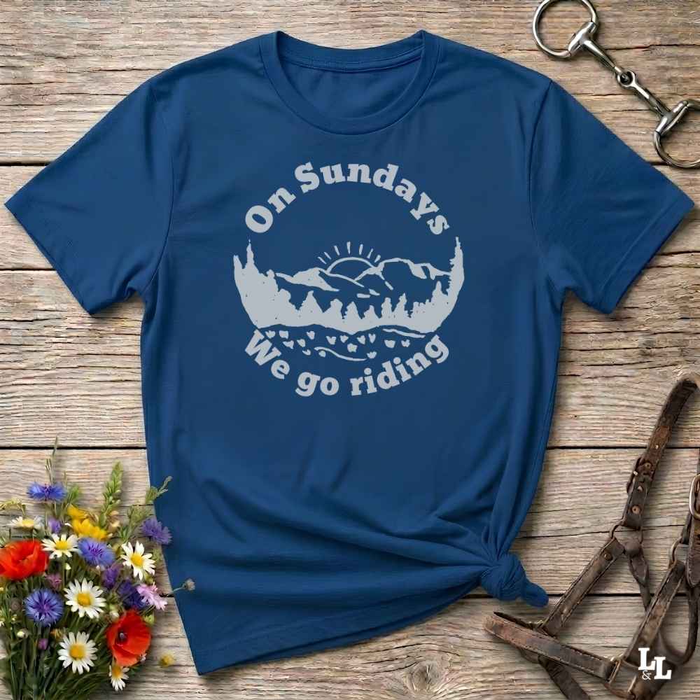 A navy blue short-sleeve t-shirt with a white graphic reading 'On Sundays We go riding' and a mountain-and-tree illustration, laid flat on a rustic wood surface with flowers and horse tack props.