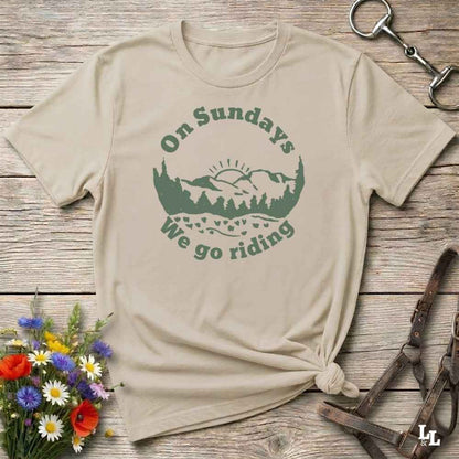A beige short-sleeve t-shirt with a green graphic reading 'On Sundays We go riding' around a mountain and tree silhouette, styled flat on a rustic wooden surface with flowers and leather tack nearby.