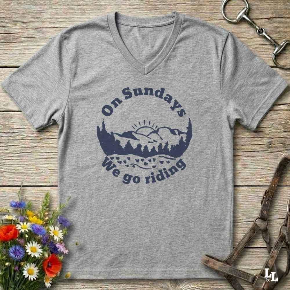 A heather gray V-neck t-shirt with a navy blue graphic reading 'On Sundays We go riding' around a mountain and tree silhouette. The shirt is laid flat on a rustic wooden surface with equestrian tack and flowers as props.