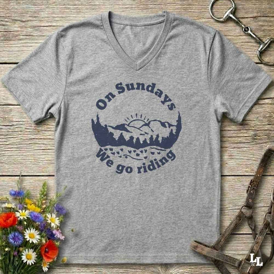 A heather gray V-neck t-shirt with a navy blue graphic reading 'On Sundays We go riding' around a mountain and tree silhouette. The shirt is laid flat on a rustic wooden surface with equestrian tack and flowers as props.
