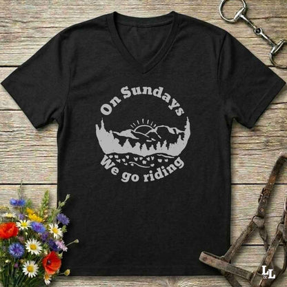 A black V-neck t-shirt with a gray graphic reading 'On Sundays We go riding' around a mountain and tree silhouette, laid flat on a rustic wood background with flowers and horse tack props.